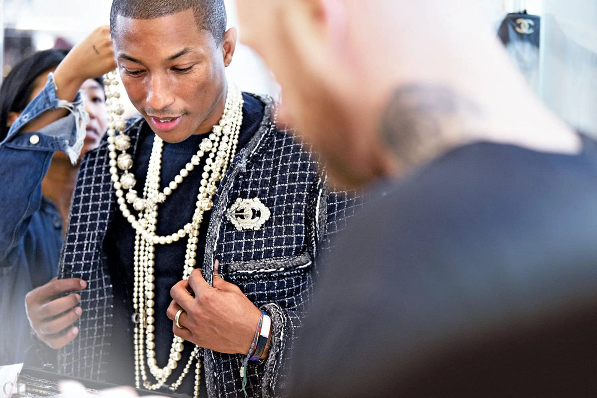 Pharrell Williams wearing a patterned jacket and multiple necklaces in an indoor setting
