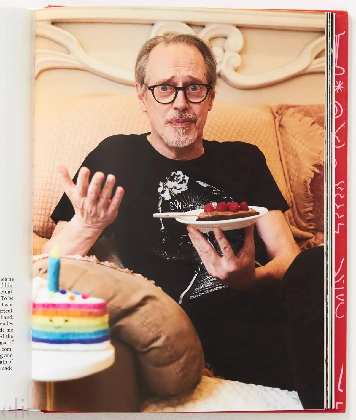 Man holding a plate with a small cake on a couch, with a rainbow cake in front