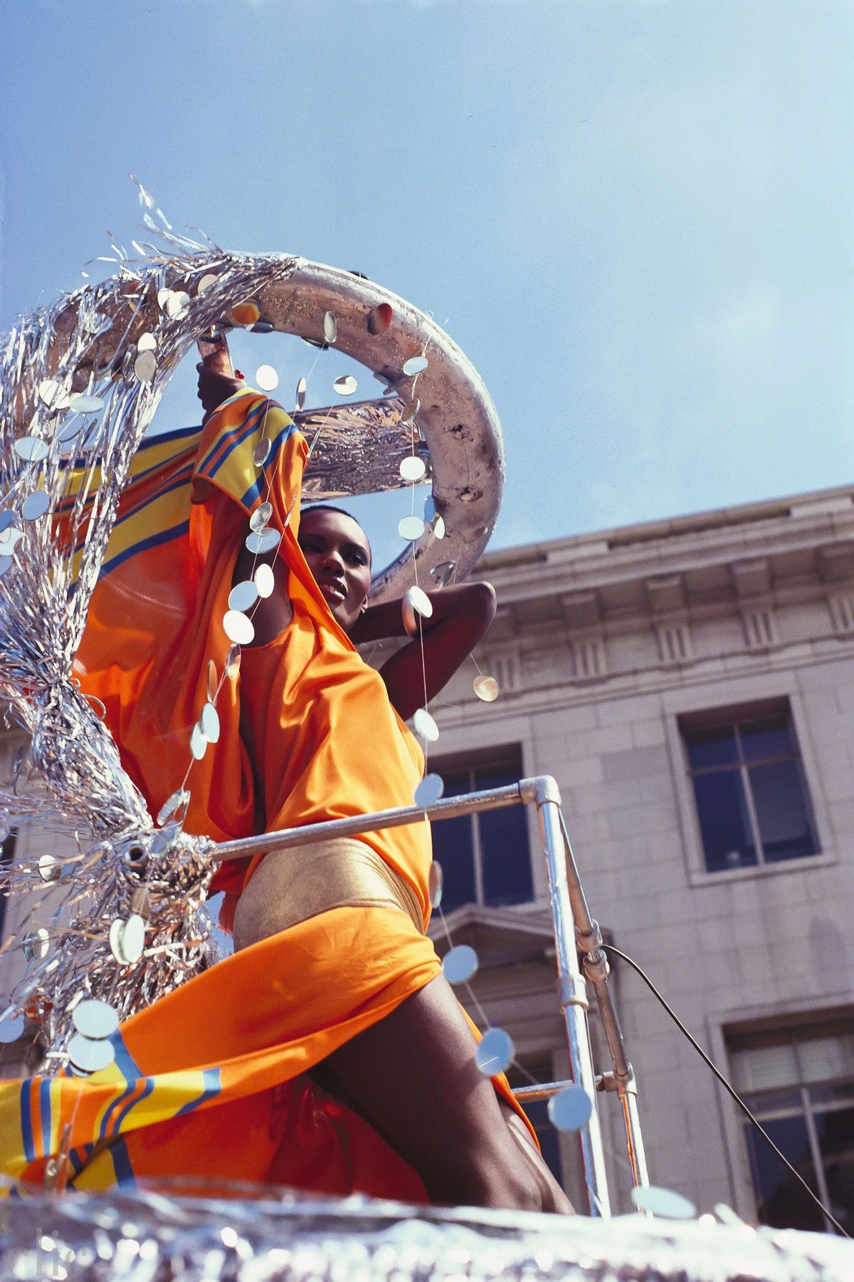 Grace Jones in an orange outfit standing on a float with decorative elements, against a clear blue sky.