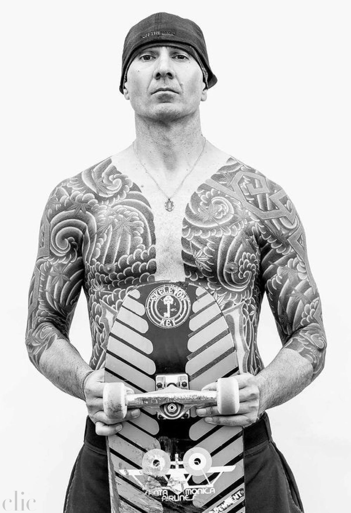 Person with extensive tattoos holding a skateboard against a white background
